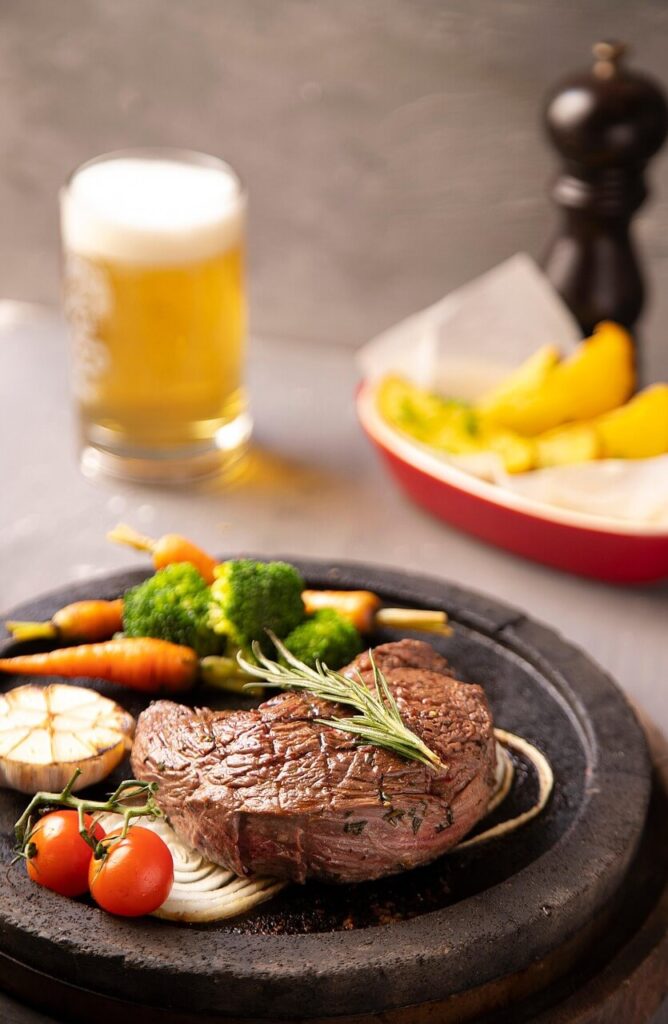Grilled Coulotte steak served with roasted vegetables, cherry tomatoes, garlic, and rosemary on a sizzling stone plate.